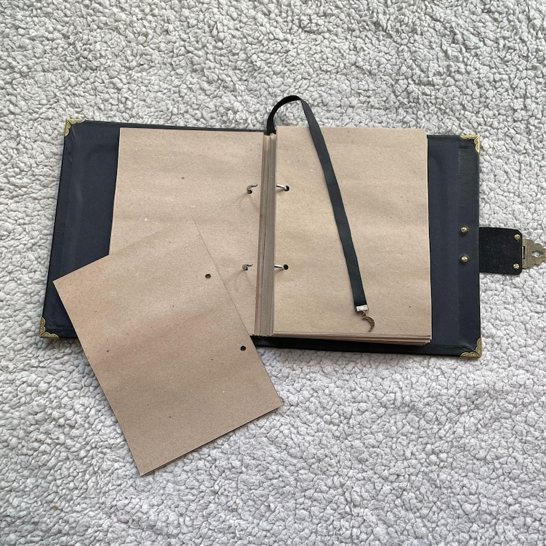 K&ouml;nnte beinhalten: Ein offenes, schwarz ledergebundenes Notizbuch mit Eckensch&uuml;tzern aus Messing und einem Verschluss. Das Notizbuch enth&auml;lt mehrere leere, hellbraune Seiten und ein schwarzes Lesezeichenband mit einem Halbmond-Anh&auml;nger. Zwei lose Seiten sind sichtbar.