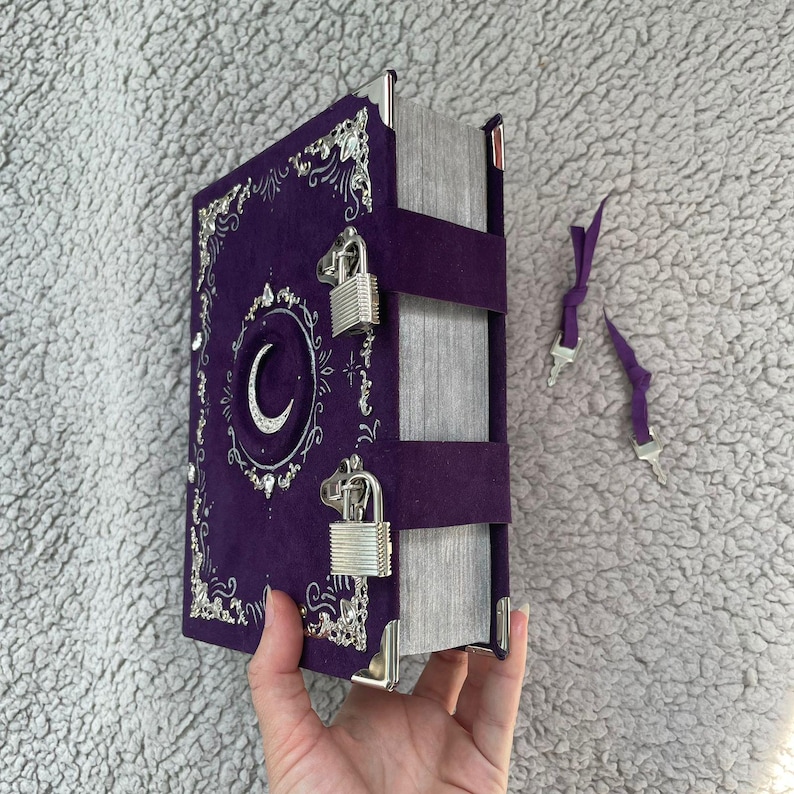 May include: A purple journal with silver detailing, a crescent moon emblem, and two silver padlocks. The journal has a velvet texture and silver corner protectors. Two purple ribbon bookmarks with silver tips are included.