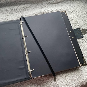 May include: Open black binder with three metal rings, a black velvet bookmark, and brass corner protectors. The binder is open to reveal black pages, and the cover has a leather clasp.