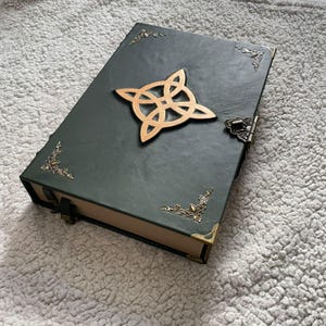 Könnte beinhalten: Ein dunkelgrünes, ledergebundenes Buch mit einem goldenen keltischen Knoten-Emblem. Das Buch hat verzierte Eckakzente und einen Verschluss. Die Kanten der Seiten sind goldfarben. Ein dekorativer und einzigartiger Artikel.