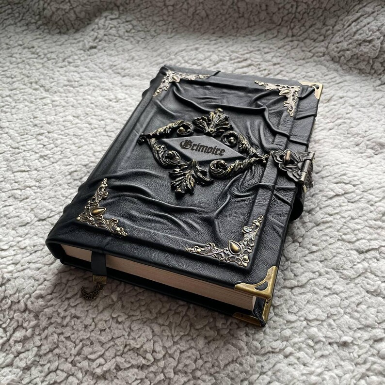 May include: A black leather-bound grimoire with ornate metal corner accents and a central decorative emblem. The word "Grimoire" is inscribed on the cover. The book has a clasp closure and a ribbon bookmark.