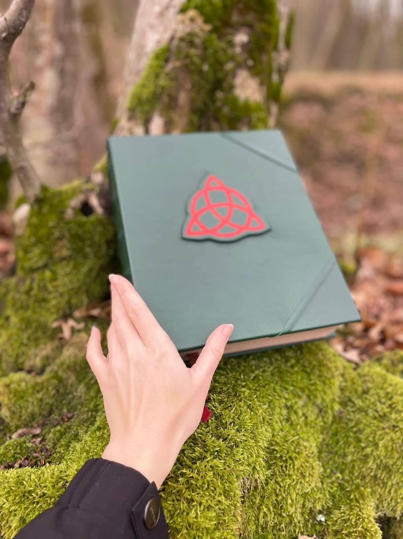 May include: A green leather-bound book with a red triquetra symbol on the cover. The book is lying on a bed of green moss.