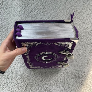 May include: A purple journal with silver-edged pages and a crescent moon design. The journal has a velvet cover with ornate silver embellishments and a clasp closure. Purple ribbon bookmarks are visible.