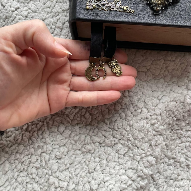 May include: Black leather journal with decorative metal corner details. Two black ribbon bookmarks with bronze charms: a crescent moon, a horseshoe, and an owl. The journal is open, showing the pages.