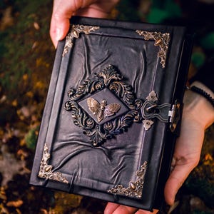 May include: A black leather-bound journal featuring a central bronze-toned moth emblem surrounded by an ornate filigree frame. The cover includes decorative metal corner protectors and a side latch closure, with a textured, creased surface finish.