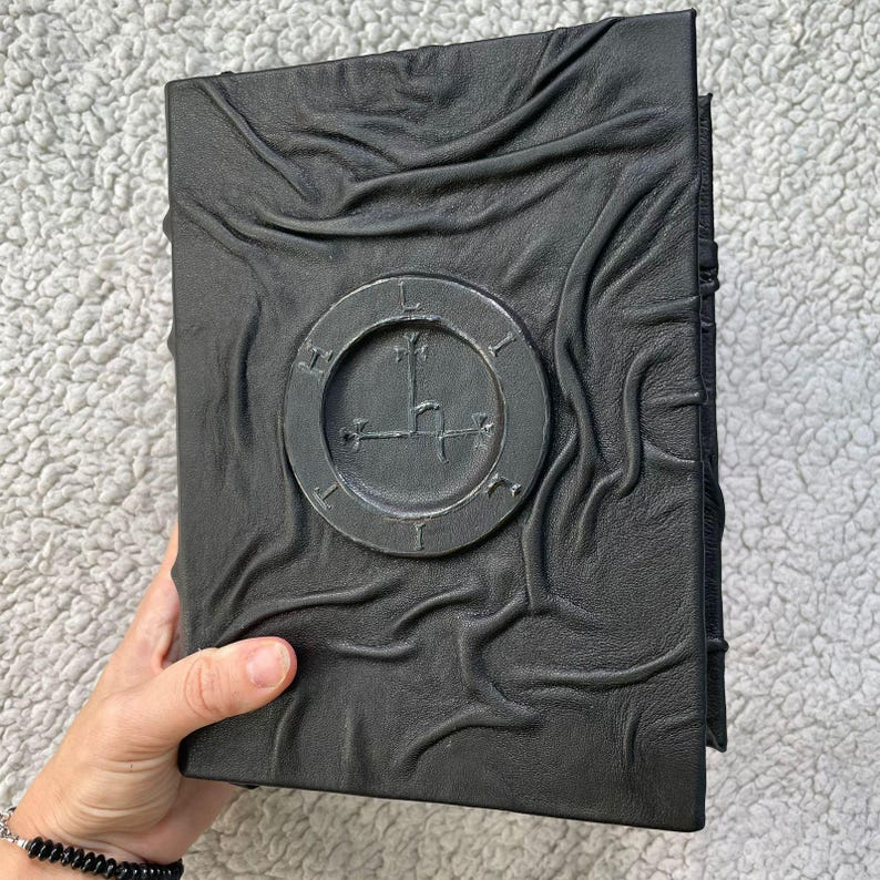 May include: A black leather-bound book with a raised circular emblem. The cover has a textured, rippled appearance. The emblem features a symbol within a circle. The book is held by a person's hand.