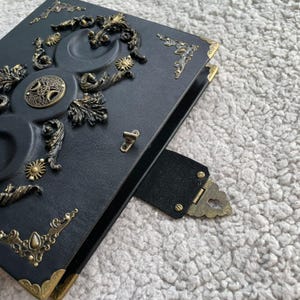 K&ouml;nnte beinhalten: Schwarzes Lederjournal mit aufwendigen Messingverzierungen. Der Einband zeigt Himmelsmotive, darunter Monde, Sonnen und dekorative Eckst&uuml;cke. Ein Messingverschluss und ein Lederriemen sichern das Journal.