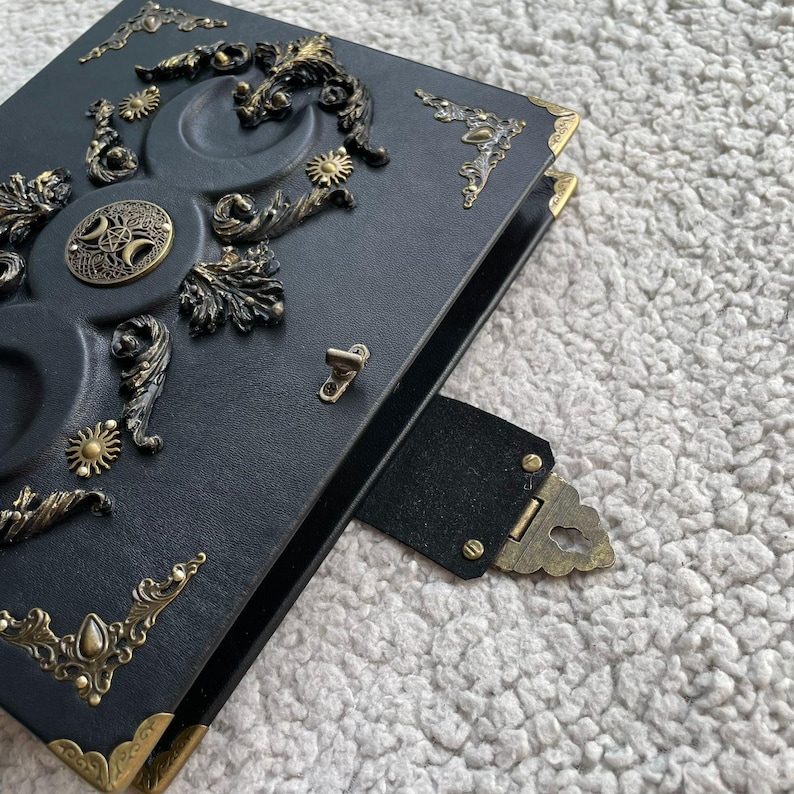 K&ouml;nnte beinhalten: Schwarzes Lederjournal mit aufwendigen Messingverzierungen. Der Einband zeigt Himmelsmotive, darunter Monde, Sonnen und dekorative Eckst&uuml;cke. Ein Messingverschluss und ein Lederriemen sichern das Journal.