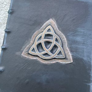 May include: A dark gray leather-bound book with a raised silver and bronze Celtic knot design. The book's spine is visible on the left side, with visible stitching. The cover has a textured appearance.