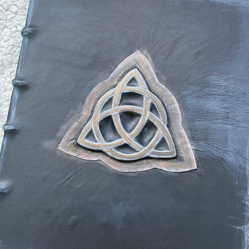 May include: A dark gray leather-bound book with a raised silver and bronze Celtic knot design. The book's spine is visible on the left side, with visible stitching. The cover has a textured appearance.