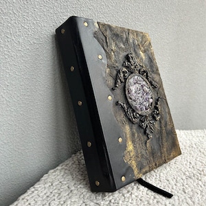 May include: A black leather-bound journal with gold accents and a decorative amethyst crystal inlay. The journal has a distressed look and a black ribbon bookmark.