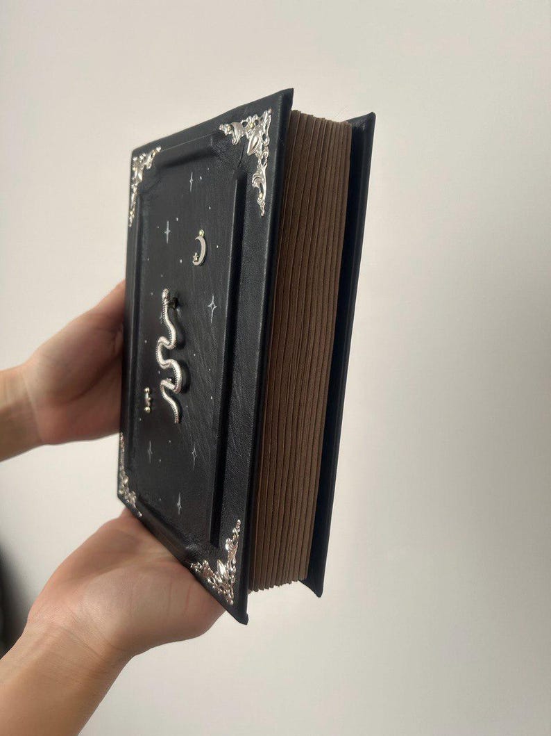 Op de afbeelding: Een zwart lederen gebonden dagboek met zilveren accenten en een slangontwerp. De omslag heeft een halve maan en sterren.