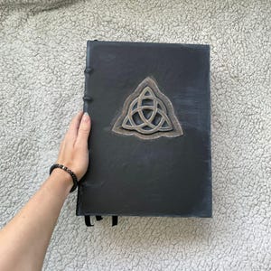 May include: A black journal with a silver Celtic knot design on the cover. The journal is bound with black thread and has a ribbon bookmark. The cover has a textured appearance.