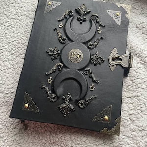 May include: Black leather journal with ornate metal embellishments. The cover features a triple moon design, stars, and decorative corner pieces. A metal clasp secures the journal.