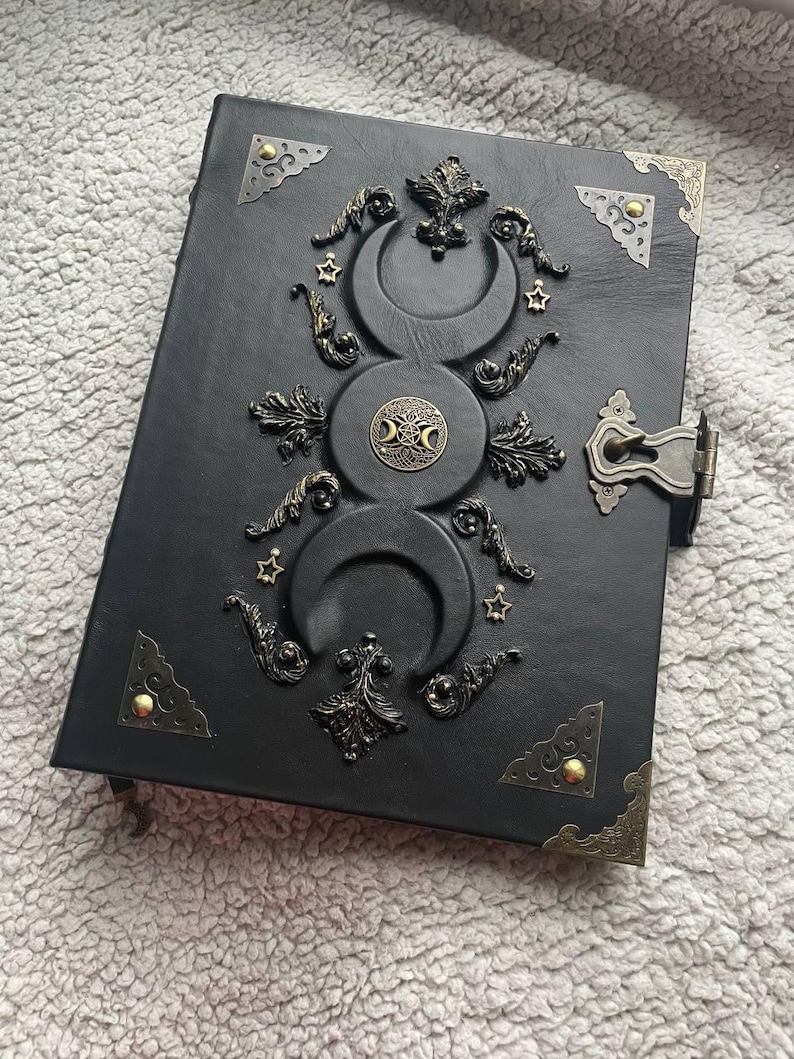 May include: Black leather journal with ornate metal embellishments. The cover features a triple moon design, stars, and decorative corner pieces. A metal clasp secures the journal.