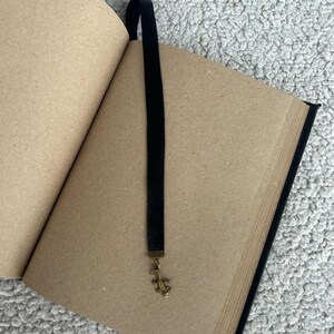 Pode incluir: Um di&aacute;rio preto com capa de papel castanho e um marcador de fita preta com um amuleto de lagarto dourado.