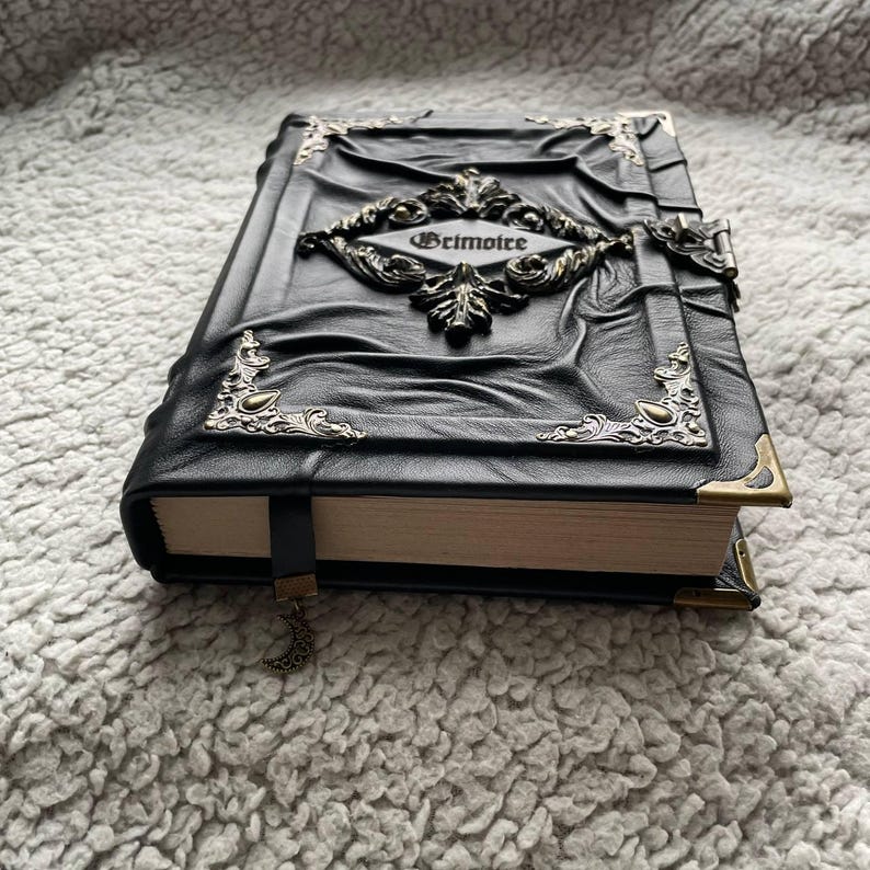 May include: A black leather-bound book with ornate metal corner accents and a central decorative emblem. The word "Grimoire" is embossed on a central plaque. A black ribbon bookmark with a crescent moon charm is visible.