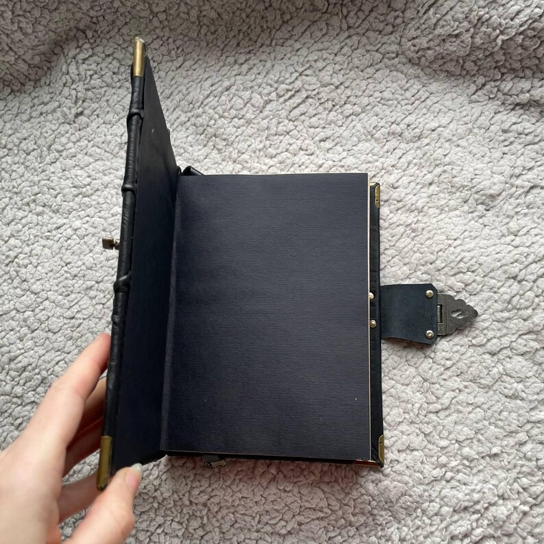 May include: Black leather journal with a clasp closure and brass corner accents. The journal is open, revealing blank black pages. The spine is stitched, and the cover is slightly textured.
