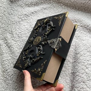 May include: A black, ornate book with gold-coloured corner accents and decorative embellishments. The cover features a central emblem and a clasp closure. The book is held open, revealing the pages inside.