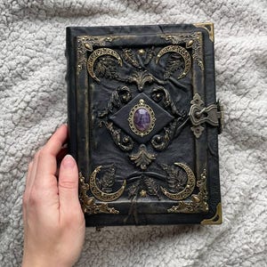 May include: Black leather journal with ornate brass corner accents and a central gemstone. The cover features embossed crescent moons, foliage, and decorative metalwork. A clasp secures the journal, which is resting on a textured, light gray surface.