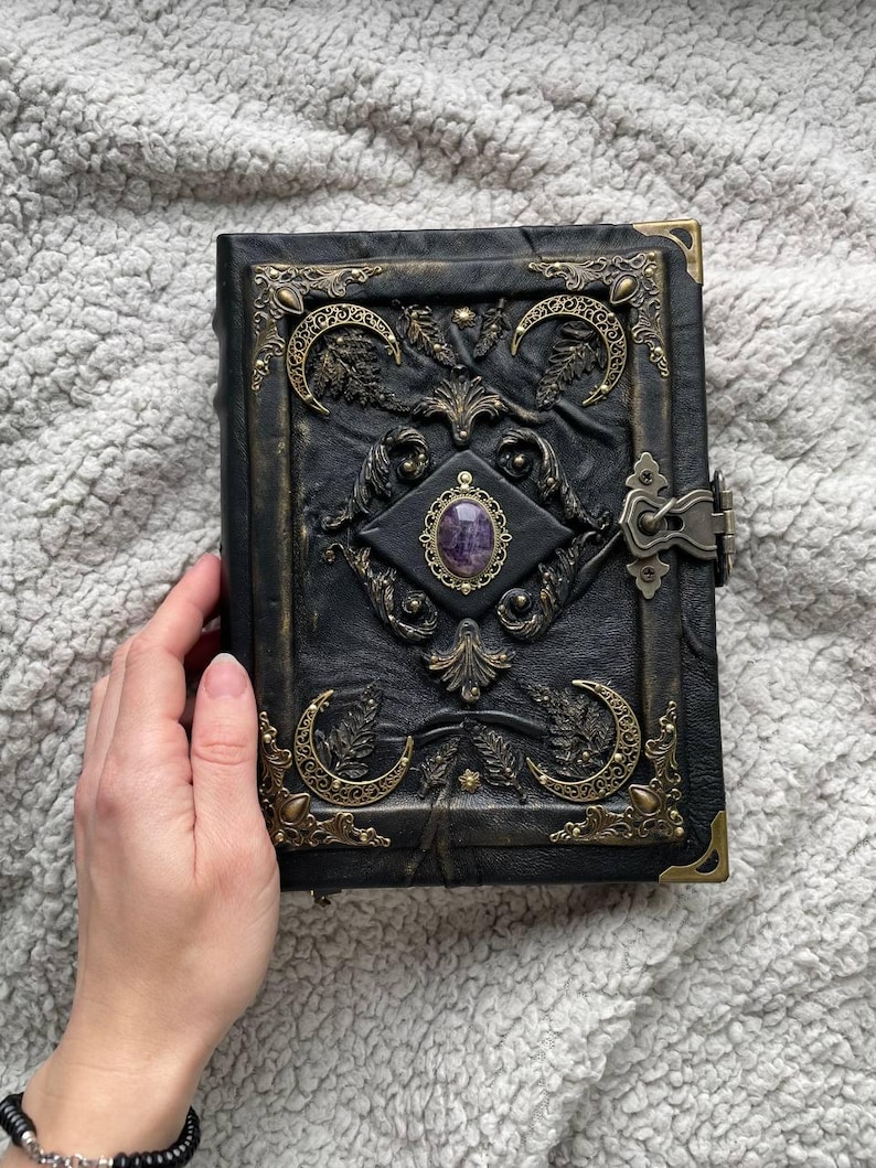 May include: Black leather journal with ornate brass corner accents and a central gemstone. The cover features embossed crescent moons, foliage, and decorative metalwork. A clasp secures the journal, which is resting on a textured, light gray surface.