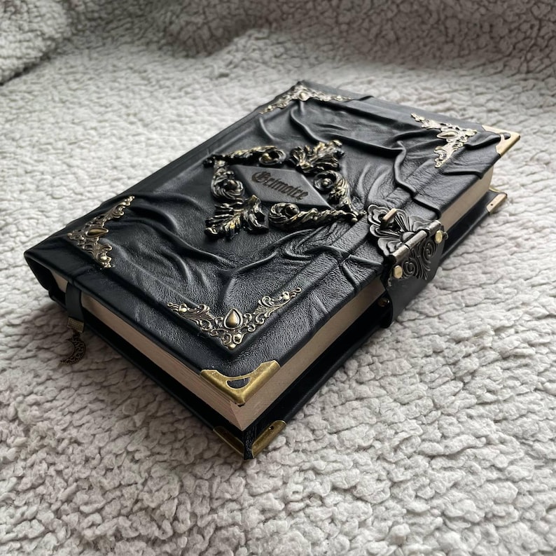 May include: A black leather-bound journal with ornate metal corner protectors and accents. The cover features embossed designs and a decorative clasp. The word "Grimoire" is embossed on the cover. The journal is closed with a leather strap.