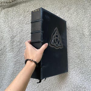 May include: A large, black book with a Celtic knot emblem on the cover. The spine has raised bands and gold-colored studs. A black ribbon bookmark hangs from the bottom. The book is held by a person.