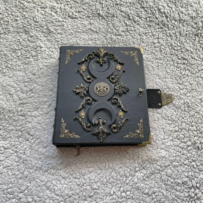 K&ouml;nnte beinhalten: Ein dunkelgraues, ledergebundenes Notizbuch mit aufwendigen goldfarbenen Metallverzierungen. Das Cover zeigt ein dreifaches Monddesign, Sonnenstrahlen und Eckakzente. Ein kleiner, schwarzer Verschluss sichert das Notizbuch.