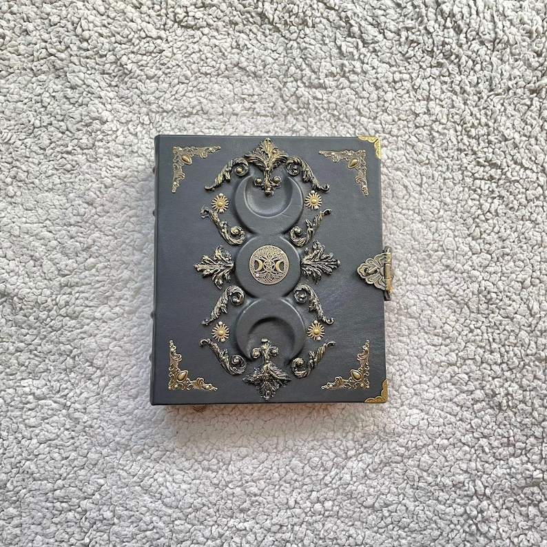 K&ouml;nnte beinhalten: Ein dunkelgraues, ledergebundenes Notizbuch mit aufwendigen goldfarbenen Metallverzierungen. Das Cover zeigt ein dreifaches Monddesign, Sonnenstrahlen und ein zentrales Emblem. Dekorative Eckst&uuml;cke und ein Verschluss vervollst&auml;ndigen das Design.