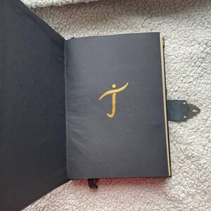 Pu&ograve; includere: Un diario nero aperto con una "J" dorata sulla copertina. Il diario &egrave; assicurato da un cinturino in pelle nera con una chiusura in metallo. Un segnalibro a nastro nero &egrave; visibile. Le pagine sono nere.