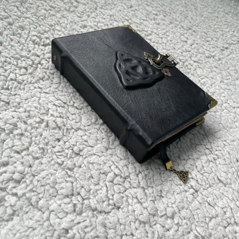 May include: Black leather journal with a Celtic knot design on the cover. The journal has a metal clasp closure and corner protectors. A decorative bookmark with a matching charm hangs from the top.