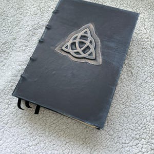 May include: A black leather-bound journal with a silver Celtic knot emblem. The journal has a textured cover and a sewn spine. Black ribbon bookmarks are visible.
