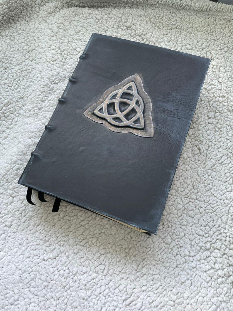 May include: A black leather-bound journal with a silver Celtic knot emblem. The journal has a textured cover and a sewn spine. Black ribbon bookmarks are visible.