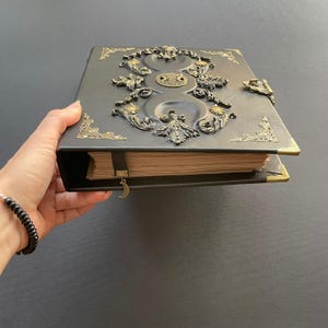 K&ouml;nnte beinhalten: Ein dunkelgraues, ledergebundenes Notizbuch mit aufwendigen, gepr&auml;gten Details. Das Cover zeigt ein zentrales Design mit Sonnen- und Mondmotiven, akzentuiert durch goldfarbene Eckst&uuml;cke und einen Verschluss. Ein goldener Halbmond-Anh&auml;nger h&auml;ngt am Lesezeichen.