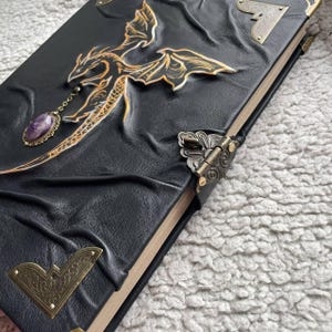 Pu&ograve; includere: Diario in pelle nera con un disegno di drago in rilievo dorato e un ciondolo con pietra viola. Il diario ha protezioni angolari in metallo e una chiusura a fermaglio. La copertina &egrave; strutturata.