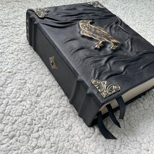 Pu&ograve; includere: Diario rilegato in pelle nera con un'aquila in rilievo e dettagli decorativi agli angoli. Il diario presenta una chiusura a fermaglio e segnalibri a nastro neri. La copertina ha un design in rilievo strutturato.