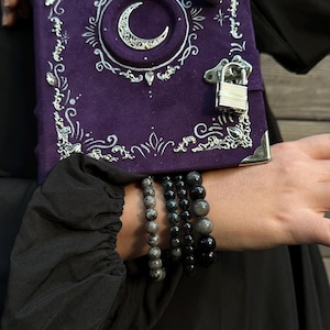 May include: A purple journal with a crescent moon design and silver detailing. The journal features a lock closure and decorative corners. Several beaded bracelets in grey and black are visible.