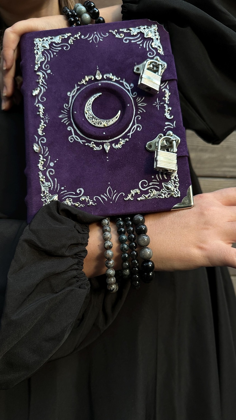 May include: A purple journal with a crescent moon design and silver detailing. The journal features a lock closure and decorative corners. Several beaded bracelets in grey and black are visible.
