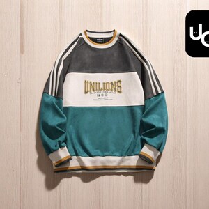 Puede incluir: Una sudadera con bloques de color en verde azulado, blanco y gris con el texto "UNILIONS" en el pecho. La sudadera tiene rayas blancas en las mangas y cuello y puños acanalados.