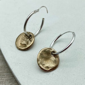 以下が含まれることがあります： 銀のフープピアスに、テクスチャのあるゴールドの円盤チャームがそれぞれぶら下がっている。