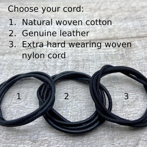 May include: Three different types of cord for jewellery making: natural woven cotton, genuine leather, and extra hard wearing woven nylon cord. The cords are black and are shown coiled on a wooden surface.
