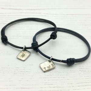 Pode incluir: Duas pulseiras de couro com pingentes de prata. Um pingente tem a letra "R" e o outro pingente tem a letra "T" com um símbolo de coração.