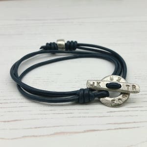May include: A dark blue leather bracelet with a silver toggle clasp. The toggle has the words "Forever" and "JKTR" engraved on it.