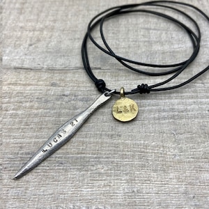 May include: A silver necklace with a long, thin pendant that says "LUCAS 21" and a round, gold pendant that says "L&K". The necklace is on a black leather cord.