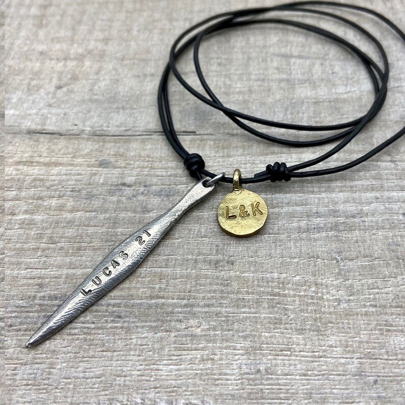 May include: A silver necklace with a long, thin pendant that says "LUCAS 21" and a round, gold pendant that says "L&K". The necklace is on a black leather cord.