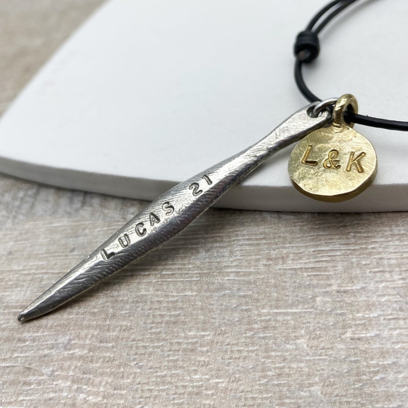 May include: A silver necklace with a long, thin pendant engraved with "LUCAS 21". The pendant is attached to a black leather cord with a gold charm that says "L&K".