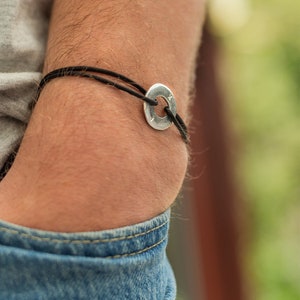 May include: A black cord bracelet with a silver circular charm. The charm has a textured surface.