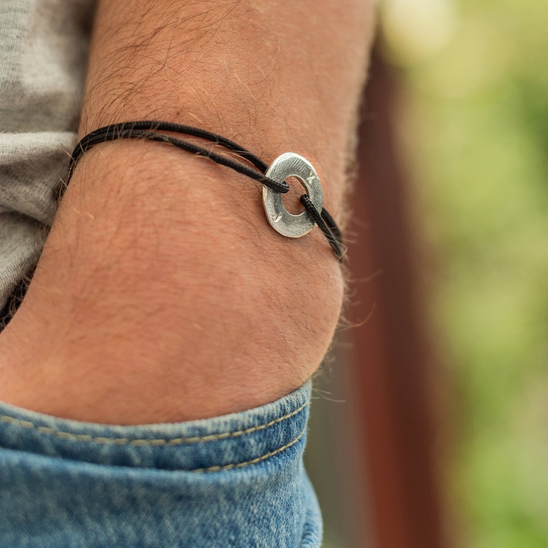 May include: A black cord bracelet with a silver circular charm. The charm has a textured surface.