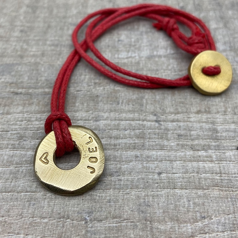 May include: A red cord necklace with two brass charms. The larger charm is a round disc with the word "JOEL" engraved on it and a small heart symbol. The smaller charm is a plain round disc.