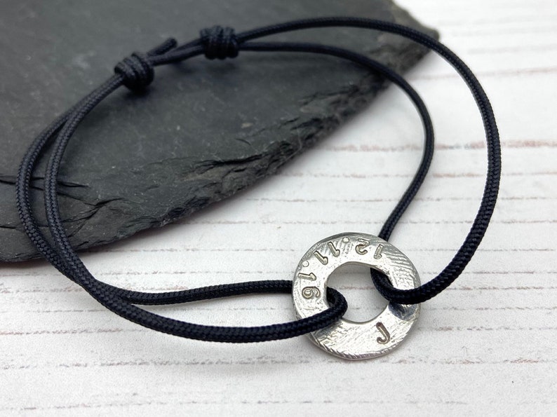 May include: A black cord bracelet with a silver metal charm. The charm is a round disc with the numbers 1-12 and the letter J engraved on it.